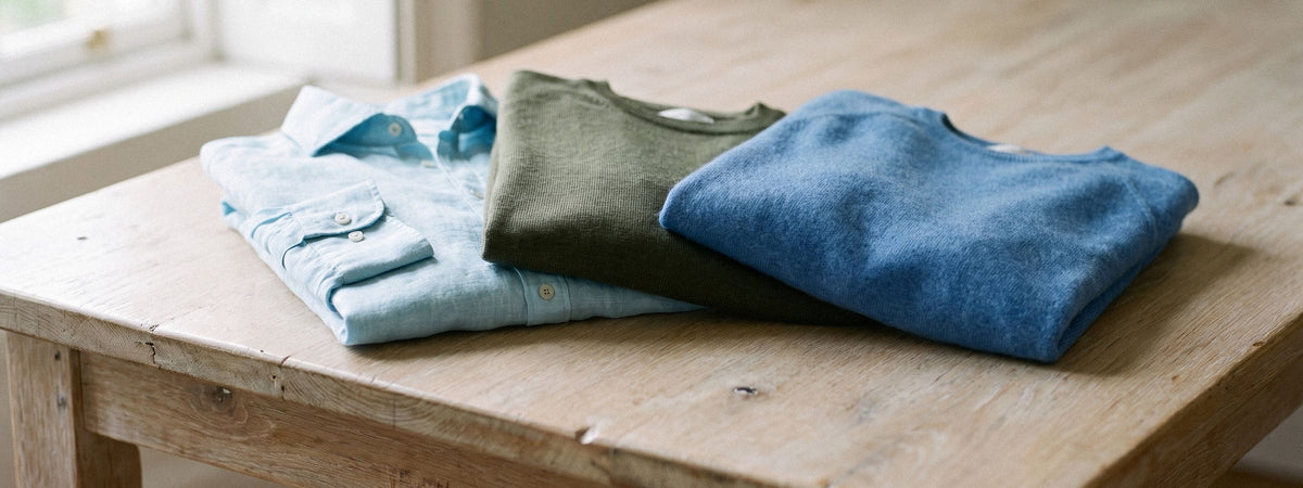 Una camicia di lino, una maglia in merino e una in cashmere piegate su un tavolo di rovere alla luce del giorno