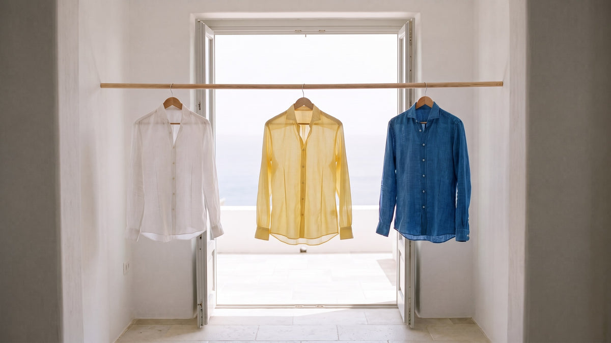 A white linen shirt, a pale yellow cotton voile shirt and a blue linen shirt hanging in a sunlit doorway, daylight passing through the voile